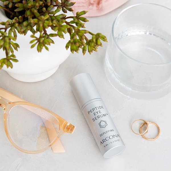 Arcona peptide eye serum beside a succulent, water, glasses, and earrings.