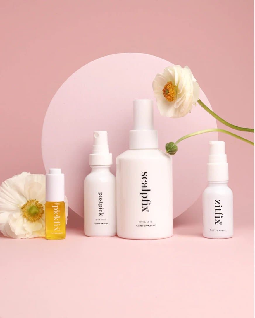 Four Carter + Jane skincare bottles with flowers on a pink background.